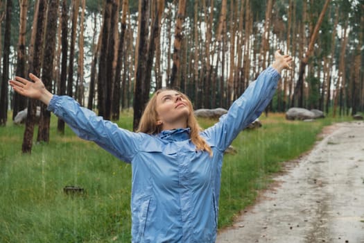Overjoyed blonde woman in blue raincoat enjoying silence natural green environment woods in the forest. Happy emotion open arms outdoors in rainy weather. Unity with nature physical mental wellbeing