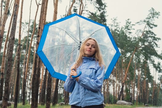 Young woman holding transparent blue umbrella outdoors in forest. Rainy weather day using umbrella. Woman with hand checking how long it will be raining. Forecast