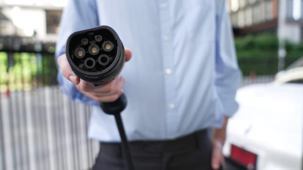 Suit-clad businessman with progressive ambition leaning pointing power cable plug at camera, charging station at city center. Electric vehicle, EV car driven by renewable energy for green environment.