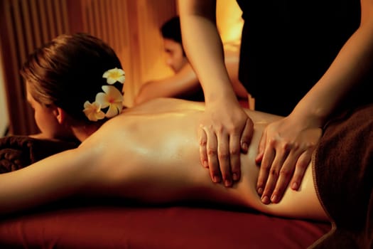 Caucasian couple customer enjoying relaxing anti-stress spa massage and pampering with beauty skin recreation leisure in warm candle lighting ambient salon spa at luxury resort or hotel. Quiescent