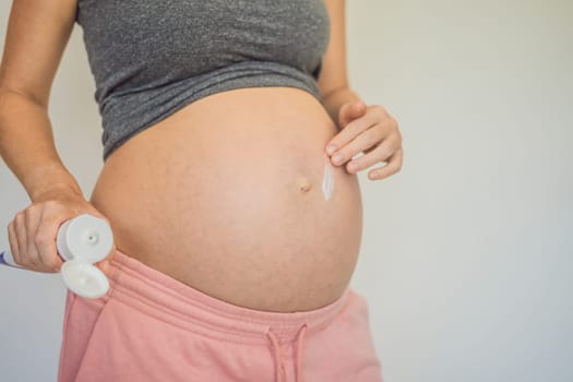 Positive young pregnant woman in comfortable homewear applying belly butter on her big tummy, woman belly.