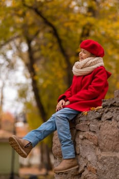 Smiling caucasian girl in a red coat and beret sits on a brick wall on a walk in autumn