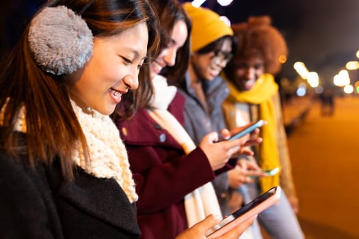 Happy young Asian female using phone with group of multiracial friends standing in a row on a cold winter night. Teens and young people addicted to technology. Copy space. Technology concept.