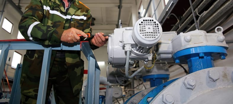 In a bustling factory setting, a skilled technician clad in a camouflage uniform diligently works on a large valve assembly. The atmosphere is filled with the hum of machinery and the scent of industrial lubricant.