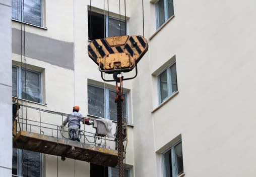 Amidst the towering structures of a bustling city, a construction worker carefully oversees renovations from a suspended platform. The crane, with its distinctive yellow and black stripes, hovers above, poised to assist in the significant task of enhancing the building's exterior.