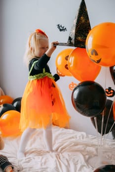Children's Halloween - a girl in a witch hat and a carnival costume with airy orange and black balloons at home. Ready to celebrate Halloween.