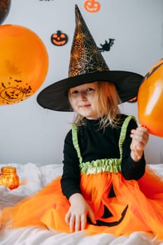 Children's Halloween - a girl in a witch hat and a carnival costume with airy orange and black balloons at home. Ready to celebrate Halloween.
