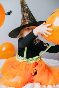Children's Halloween - a girl in a witch hat and a carnival costume with airy orange and black balloons at home. Ready to celebrate Halloween.
