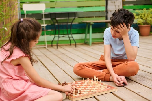 Young boy and girl engaged in a chess game while sitting on a wooden deck in a backyard, showing concentration and strategy.
