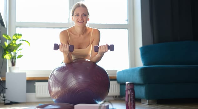 Woman doing exercises with dumbbells on a ball at home, sunny, close-up. Ball resistance training for women over 30