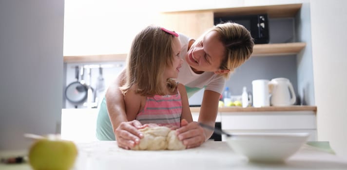 Mom and daughter knead dough together on the table, close-up, blurry. Woman teaches little girl to cook at home in the kitchen