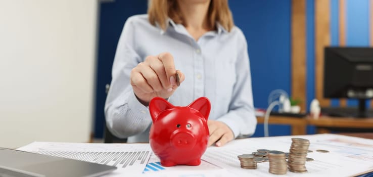 A woman in the office puts a coin in a red piggy bank, close-up. Capitalization of income, saving money, investments
