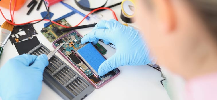 Hands in blue gloves with tweezers are repairing the battery from a smartphone, close-up. Mobile phone repair service