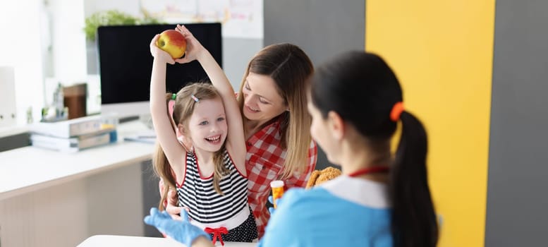 A little girl with her mother at the doctor rejoices at an apple, close-up. Consultation with a pediatric nutritionist, natural and synthetic vitamins
