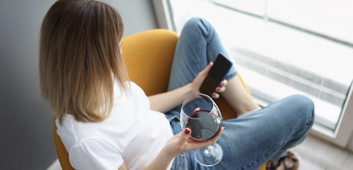 A woman at the window holds a glass of red wine and a smartphone, top view, close-up. Loneliness, stress relief, self-isolation
