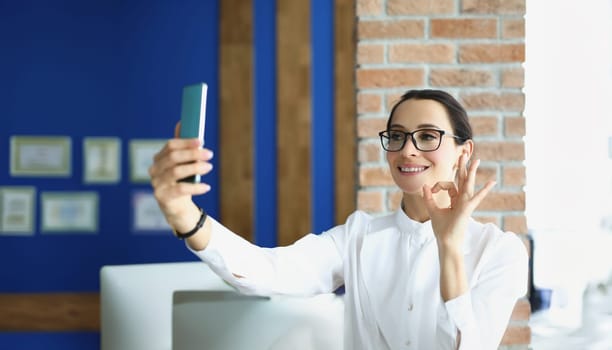 A woman in the office speaks via video calling on a smartphone, showing ok gesture close-up. Stylish businesswoman