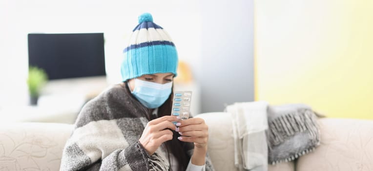 Sick woman in a mask sits in a blanket on the couch, high temperature, close-up. Medicines to treat the virus