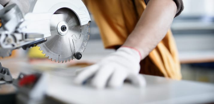 Male hands in gloves next to a circular saw, close-up, blurry. Woodworking workshop, sawing boards, carpenter equipment