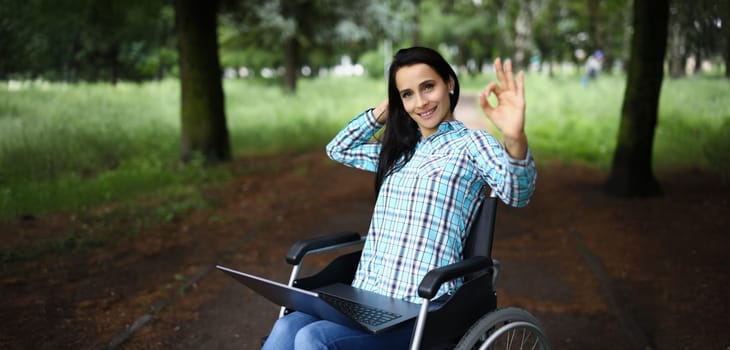 A young disabled woman sits in a wheelchair in the park with a laptop and shows the gesture ok. Psychological assistance after trauma