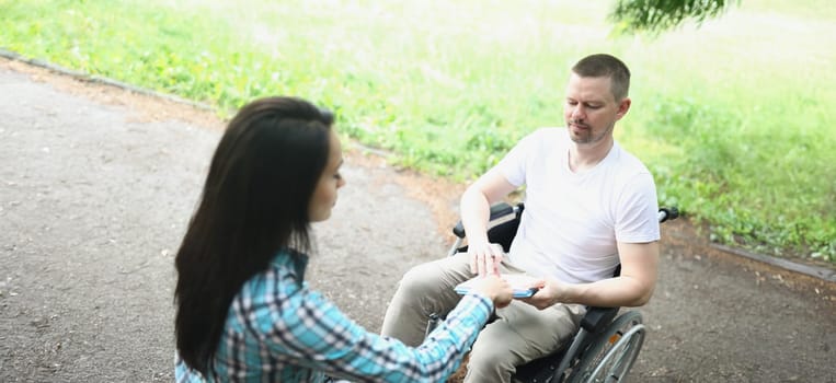 A man in a wheelchair in the park makes notes. Woman talking to a disabled person on the street. Rehabilitation after trauma