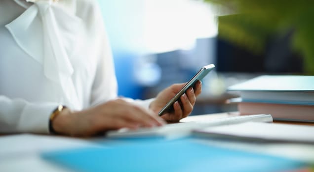 A woman works at a computer and holds a smartphone, close-up, blurry. Digital devices in the workplace, accounting department