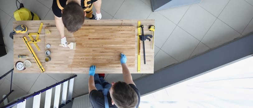Male carpenters apply varnish to a wooden surface, close-up, top view. Lacquered laminate, workshop, furniture renovation services