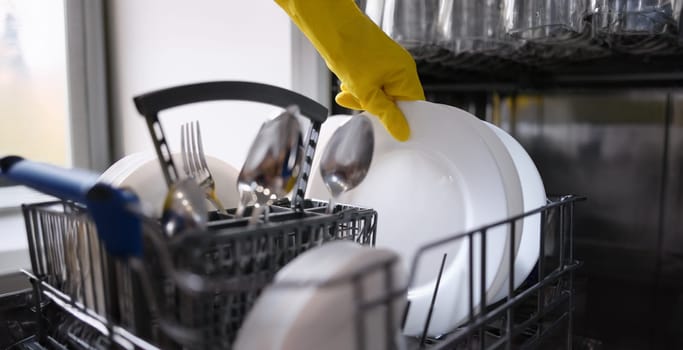 A hand in a yellow glove pulls clean dishes out of the dishwasher. Household appliances for home, kitchen utensils