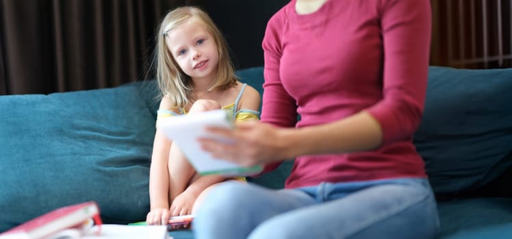 A little girl with her mother are sitting at home on the couch with books, close-up. Parent checks the child's school assignments