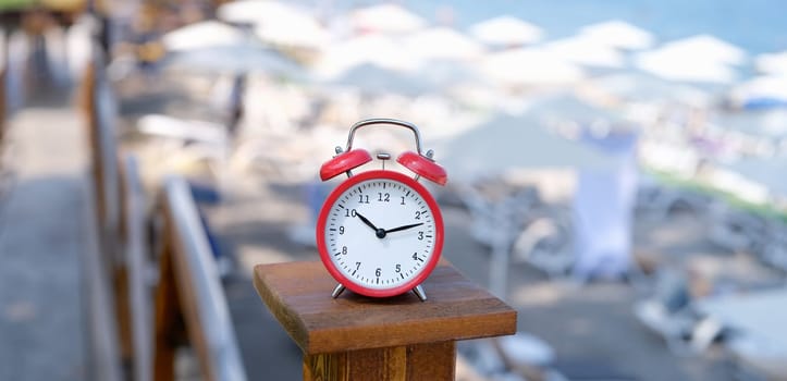 Red alarm clock on a wooden stand, against the background of beach umbrellas, close-up, blurry. Safe tanning time concept