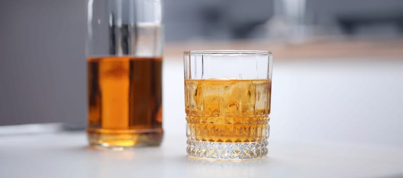 Brown alcoholic cocktail with ice in a wide glass, close-up. Incomplete whiskey bottle and glass on gray table background