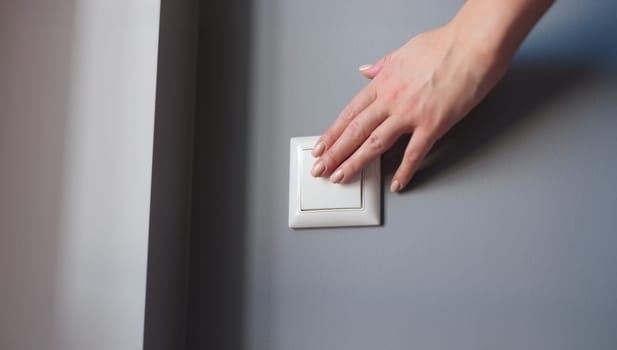 A woman's hand presses the switch on the wall, close-up. White light switch on gray wall background, checking electricity