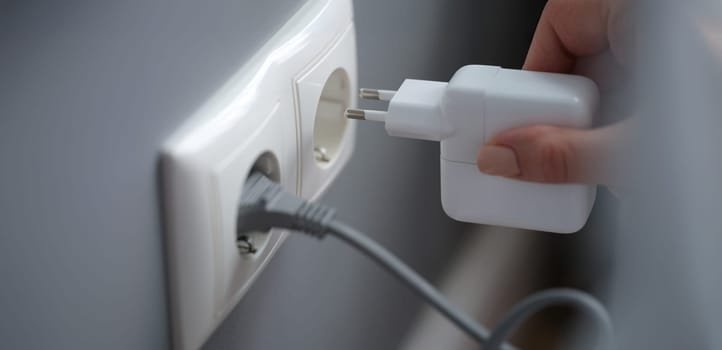 Female fingers plug the adapter into an outlet, close-up. White socket on gray wall background, electrician in apartment