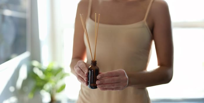 Woman holding diffuser with bamboo sticks, sunny, close-up. Aromatherapy at home, perfume, relaxation spa