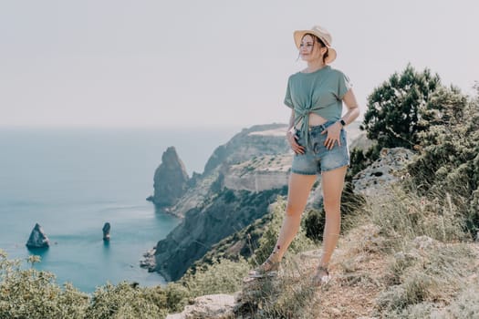 A woman stands on a cliff overlooking the ocean, wearing a hat and casual clothes, enjoying the beautiful view.