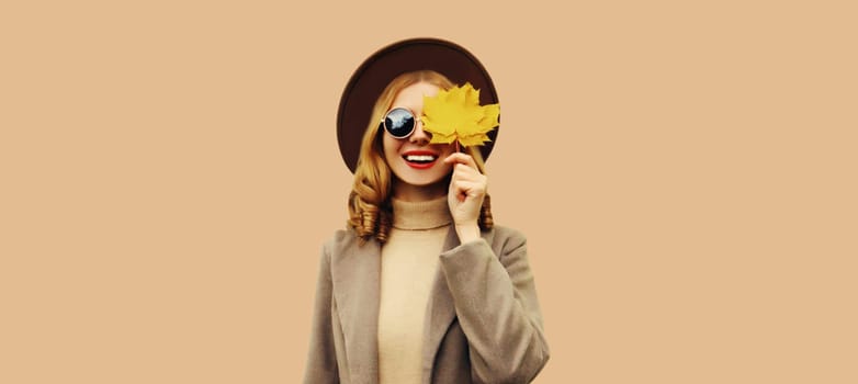 Autumn color style outfit, portrait of beautiful smiling young woman with yellow maple leaves wearing round hat, coat on brown background