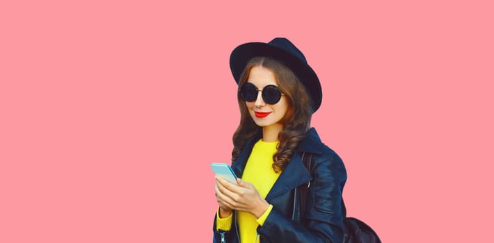 Stylish modern young woman holds mobile phone wearing black round hat on pink studio background