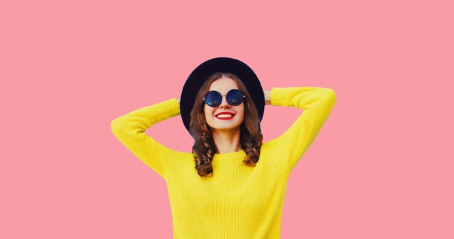 Portrait of stylish happy young woman wearing black round hat, yellow sweater on pink background