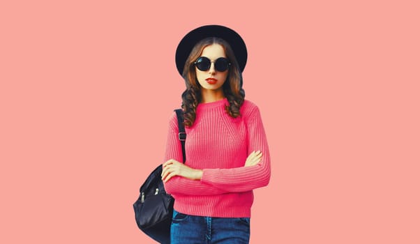 Modern young woman posing wearing sweater, black round hat and backpack on pink studio background
