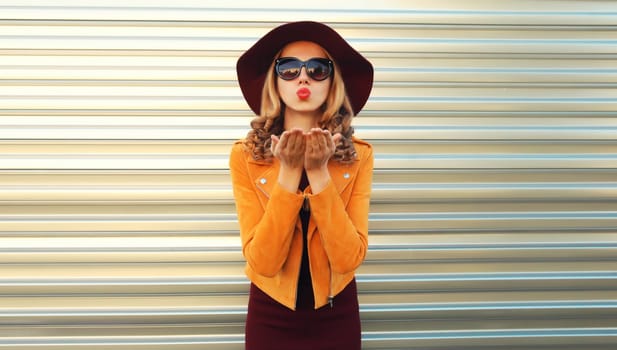 Portrait of stylish young woman posing blowing her lips sends sweet air kiss wearing sunglasses, round hat and jacket on gray background