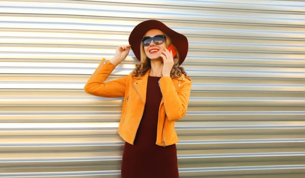 Stylish happy woman calling on smartphone looking away wearing orange jacket, hat and sunglasses on gray background