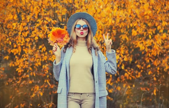 Autumn portrait of stylish beautiful young woman model with yellow maple leaves blowing her lips sends sweet kiss wearing round hat and coat in the park