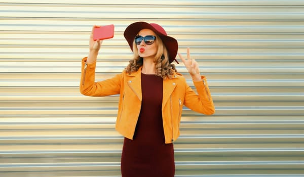 Autumn style outfit, stylish elegant woman posing taking selfie with mobile phone wearing hat, orange jacket and sunglasses on gray background