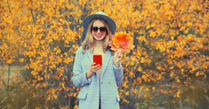 Autumn style outfit, beautiful smiling young woman model with mobile phone and yellow maple leaves wearing round hat and coat in the park