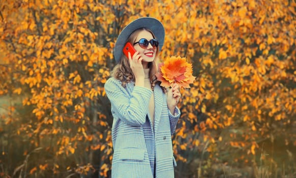 Autumn portrait of happy smiling young woman with yellow maple leaves calling on mobile phone looking away wearing round hat and coat in the park