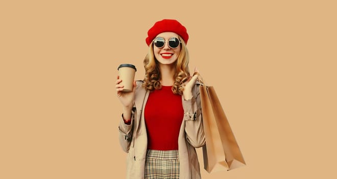 Autumn style outfit, portrait of beautiful happy smiling young woman with shopping bags holds cup of coffee wearing red french beret hat, gray coat jacket on beige studio background