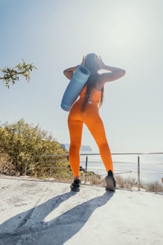 A woman in a vibrant orange workout outfit stands with her back to the camera, carrying a yoga mat and looking out at the scenic view, enjoying the beautiful day.