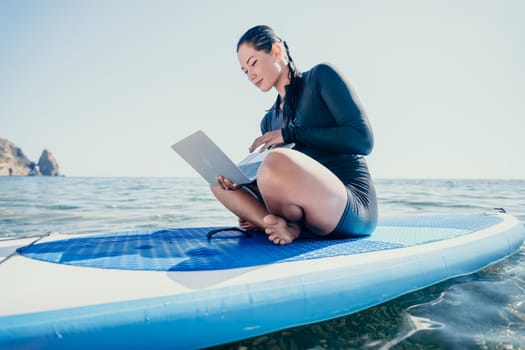 Digital nomad, woman in the hat, a business woman with a laptop sits on the rocks by the sea during sunset, makes a business transaction online from a distance. Freelance, remote work on vacation.