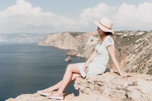A young woman in a straw hat and a light-green dress sits on a cliff overlooking the ocean and a coastline, her expression thoughtful and contemplative.