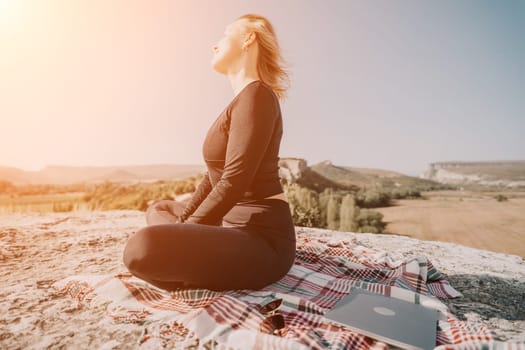 A woman in black clothing sits cross-legged on a mountaintop with a blanket and a laptop beside her, looking peaceful and serene.