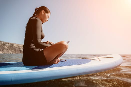 Digital nomad, woman in the hat, a business woman with a laptop sits on the rocks by the sea during sunset, makes a business transaction online from a distance. Freelance, remote work on vacation.